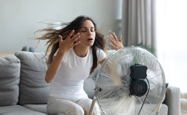 In living room without air-conditioner tired from summer heat young woman turned on floor ventilator waving her hands to cool herself, female sitting on couch suffers from unbearable too hot weather